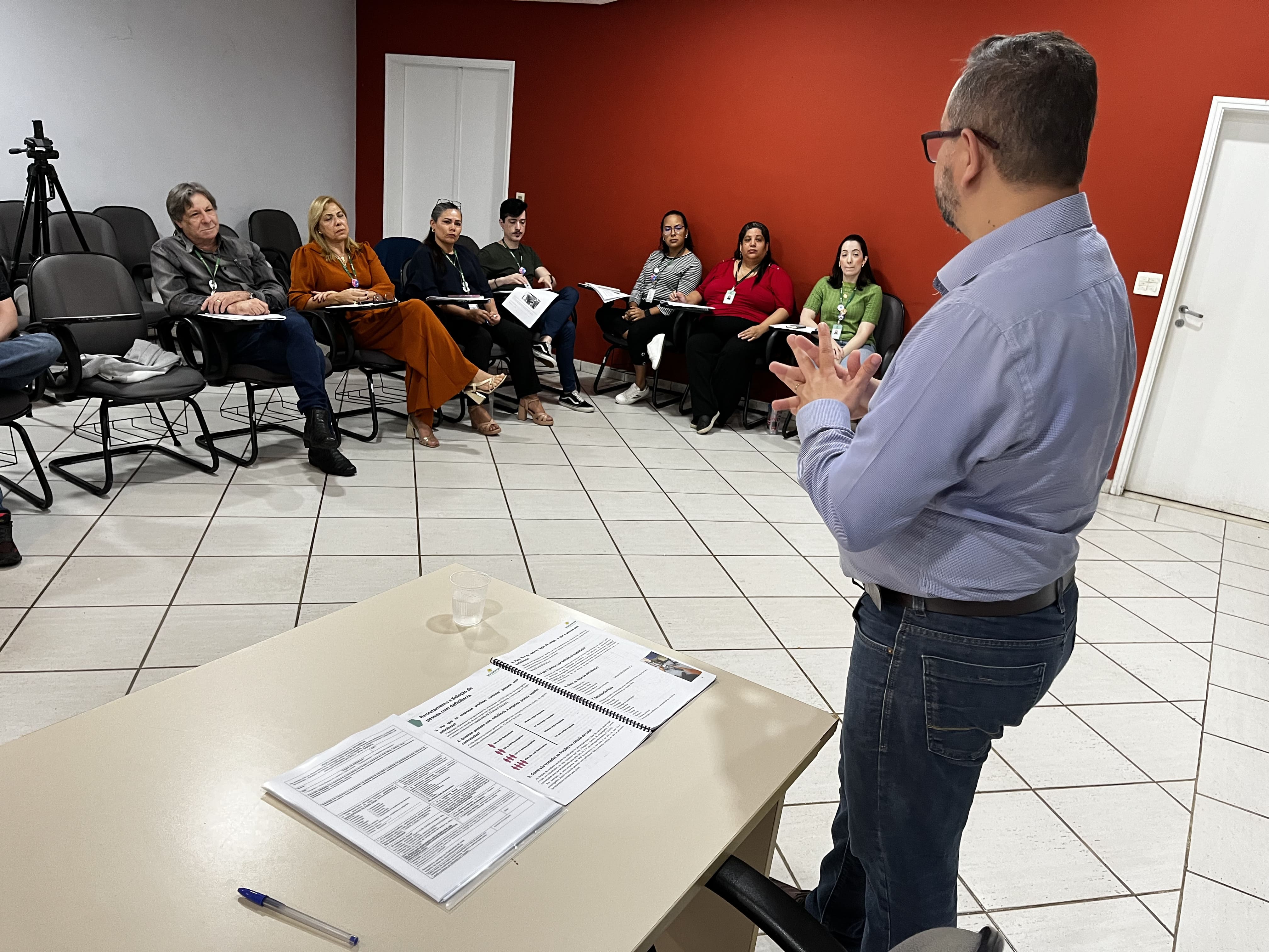 Ao fundo, sentados, os alunos do curso de Atendimento de
									qualidade para pessoas com deficiência observam com atenção o
									instrutor Fábio ( homem branco de cabelos e barba grisalhos vestindo
									uma camisa social azul  ) que aparece em primeiro plano de frente para
									os alunos. Na mesa, ao seu lado, está  a apostila do curso.