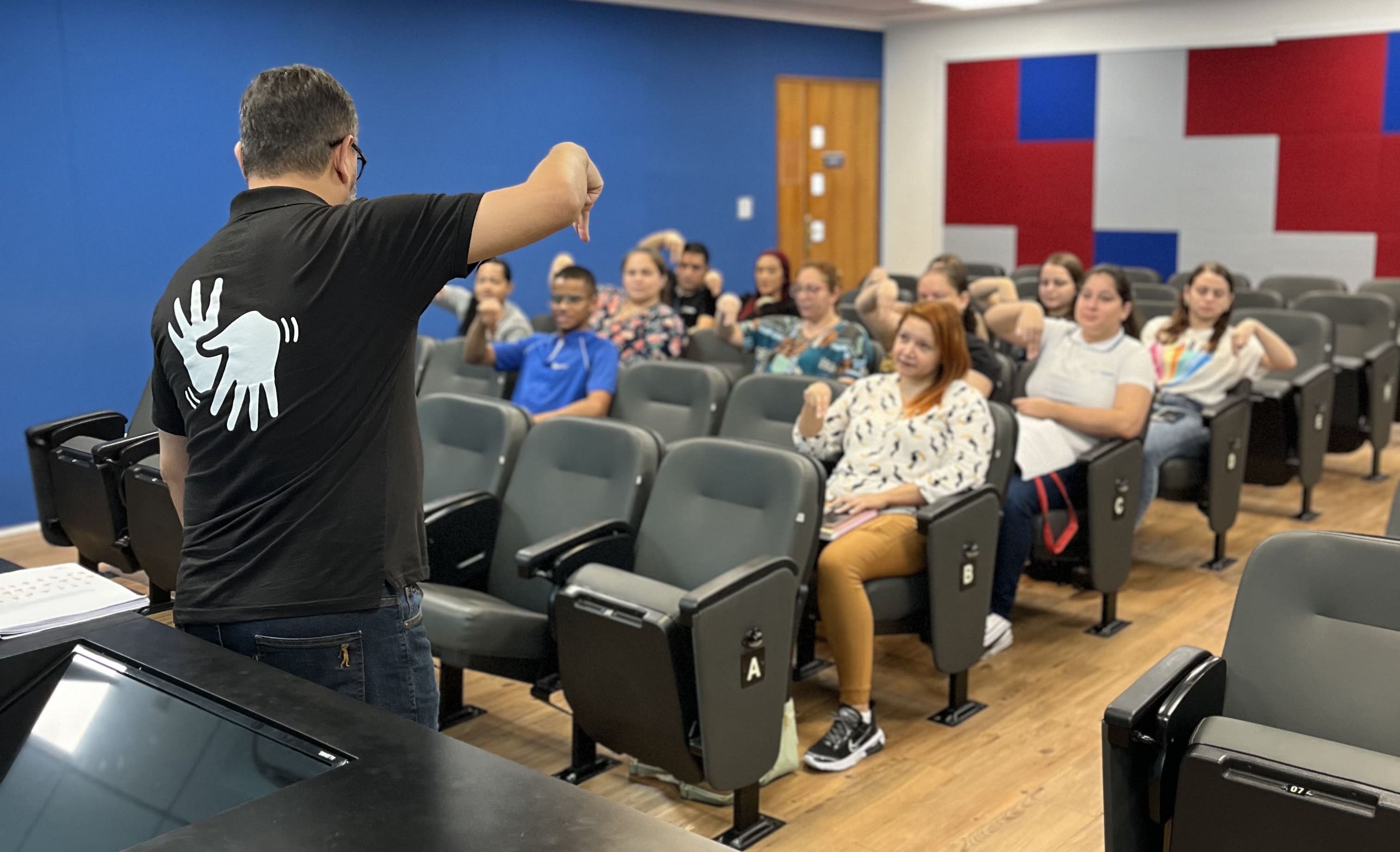 Imagem colorida. 1 homem aparece de costas para a
                                            foto realizando um sinal em Libras. Ao fundo, os alunos,
                                            sentados nas cadeiras do auditório de reuniões, repetem o
                                            sinal que o homem a sua frente faz.