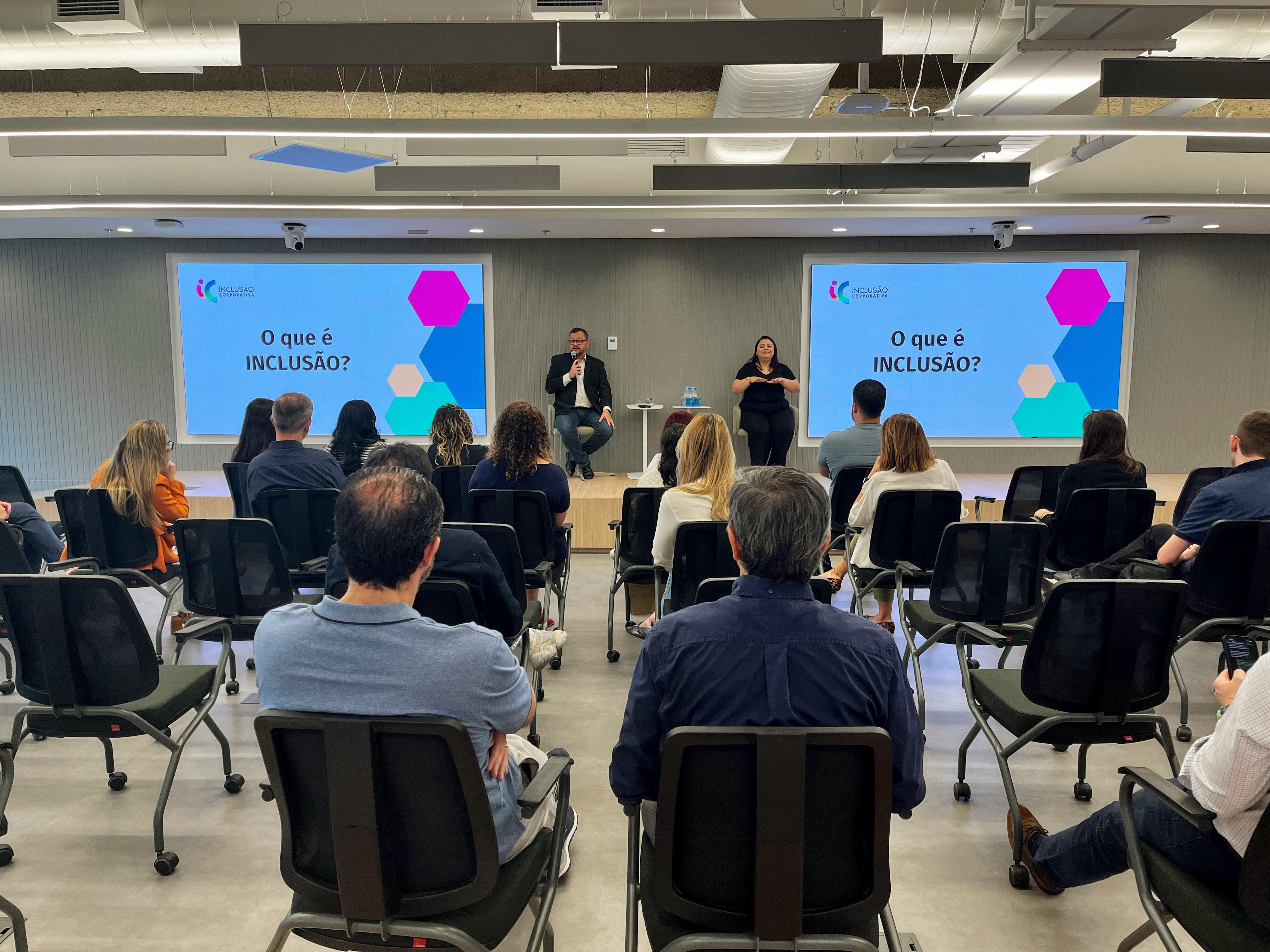 Foto colorida.  Aproximadamente 15
								pessoas sentadas de costas em uma sala
								de reuniões e palestras. No palco, ao
								fundo, o palestrante Fábio (homem
								branco usando um blaser preto) ministra a
								palestra ao lado da intérprete de Libras
								(mulher branca de cabelos longos e
								pretos). Nas paredes do palco existem
								dois televisores, apresentando slides
								escritos “ O que é Inclusão?”