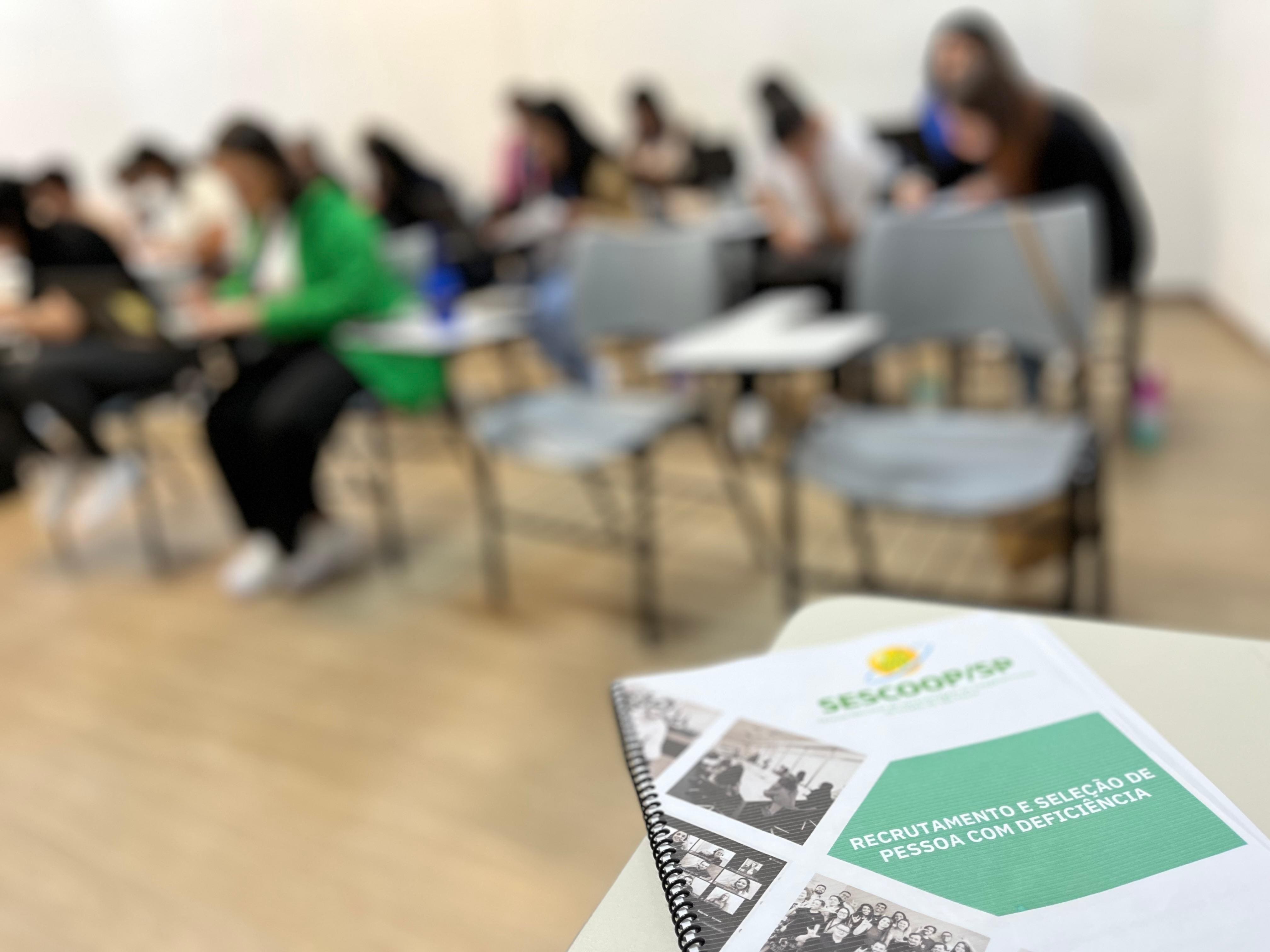 Ao fundo, com efeito de ‘desfoque’,
								os alunos do curso de Recrutamento e
								Seleção de pessoas com Deficiência
								aparecem sentados em carteiras. Em
								primeiro plano, sem ‘desfoque’, está a
								apostila do curso de Recrutamento e
								Seleção de Pessoas com Deficiência.