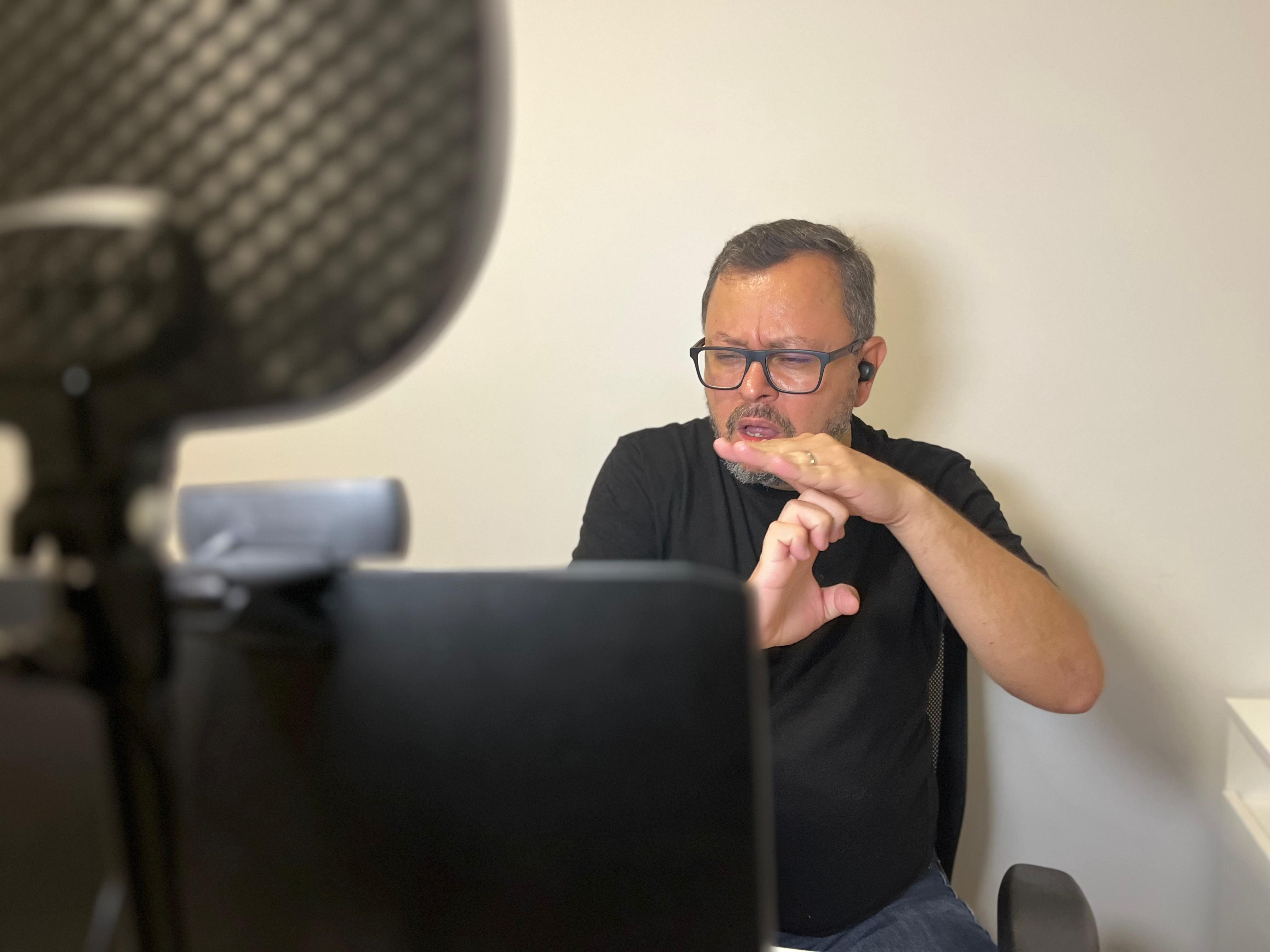 Homem branco, de óculos de grau e camiseta preta,
                                        realizando uma interpretação de Libras online. Em primeiro plano
                                        é possível ver parte do computador e da “ring ligth”  em frente ao
                                        intérprete.
