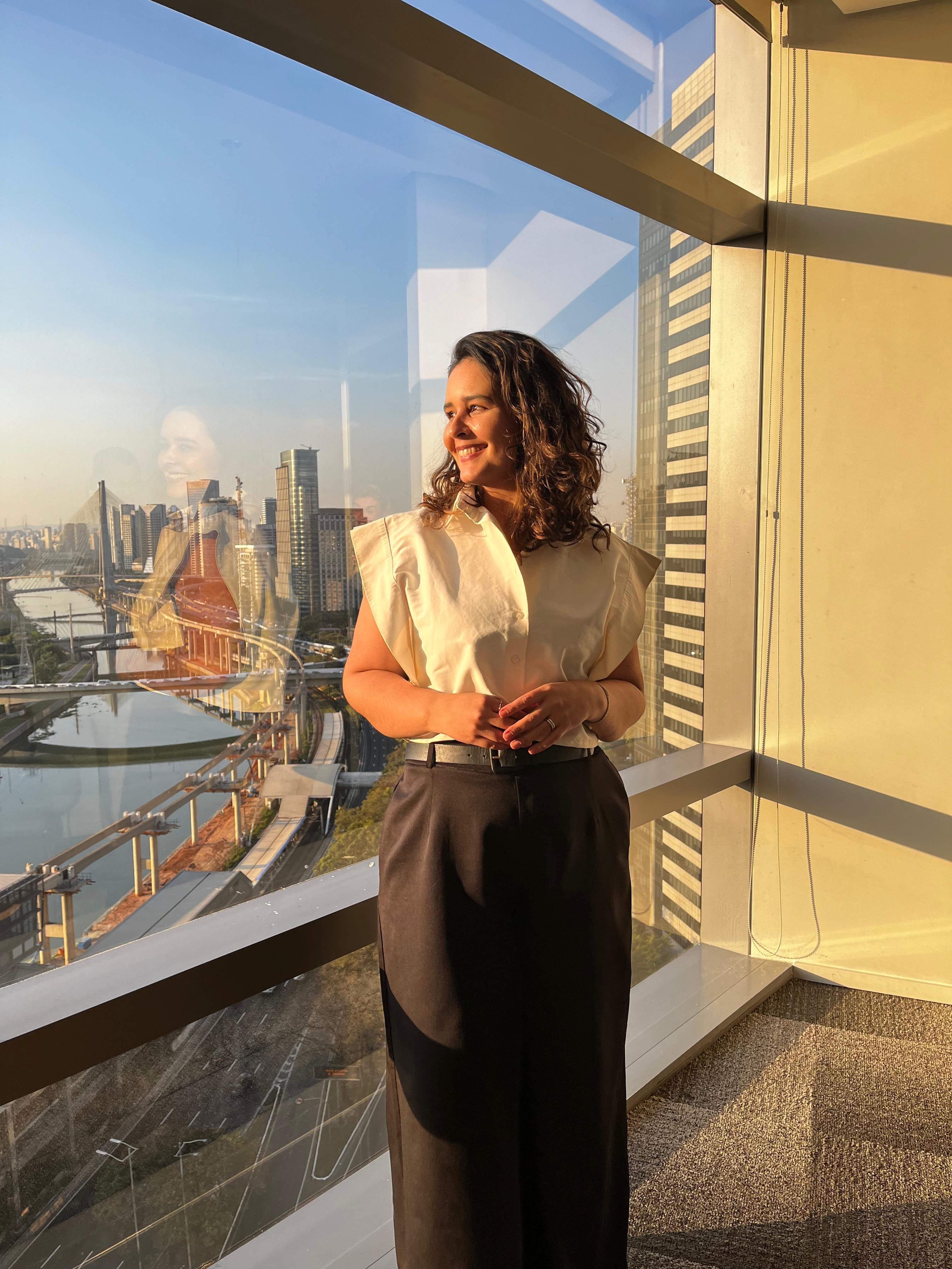 Mulher branca de cabelos castanhos na altura dos ombros. Ela veste uma camisa social branca e uma calça preta. Ao fundo, uma grande janela com vista para o rio pinheiros e prédios ao seu redor.