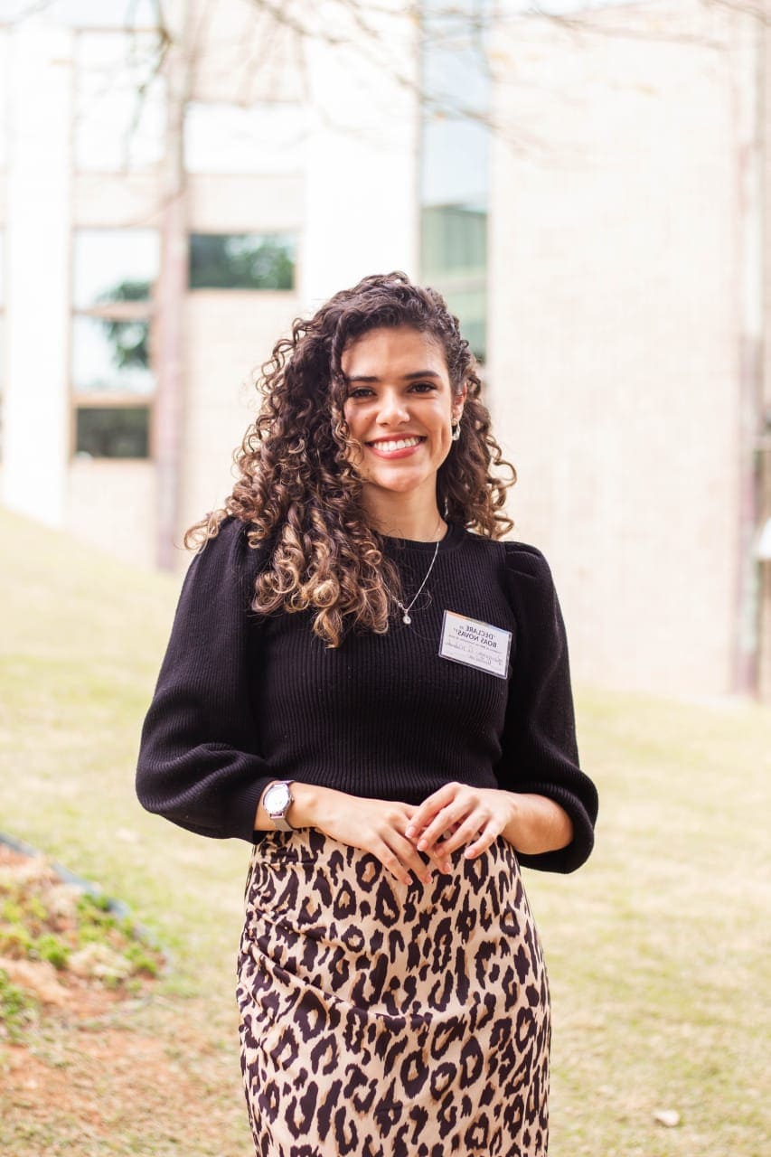 Mulher branca de cabelos cacheados abaixo dos ombros. Ela veste uma blusa meia estação na cor preta e uma saia lápis com estampa de “onçinha”. Ao fundo, parte de um edifício.
